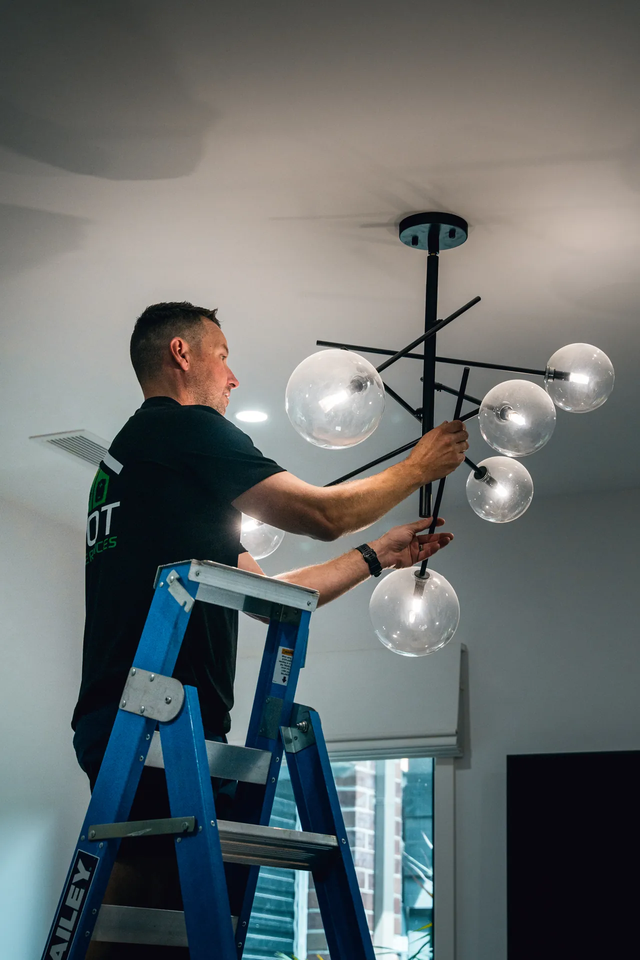 Electrician on a ladder fitting a modern multi-globe pendant light to a living room ceiling