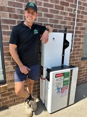 Dan Morgan standing beside a newly installed home battery and inverter on a brick wall