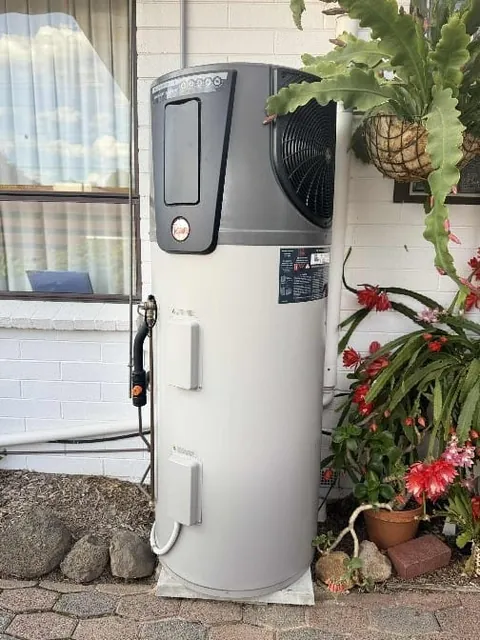 Heat pump hot water unit installed outdoors beside a brick wall and garden