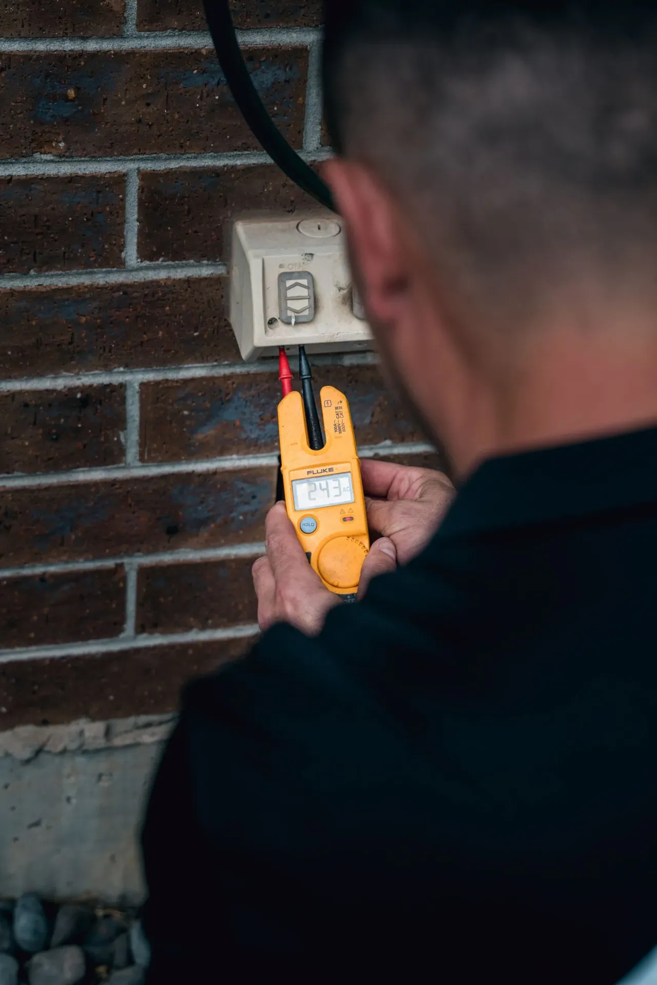 Electrician testing a weatherproof external power point on a brick wall with a voltage tester